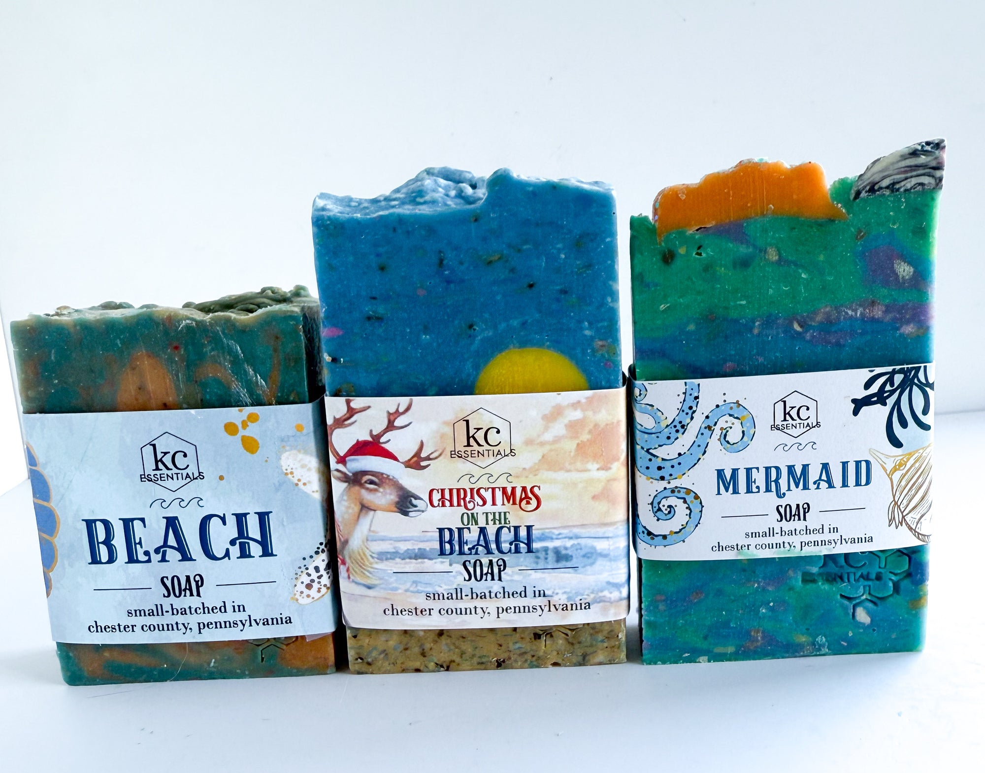 Three bars of soap with labels indicating 'Beach Soap', 'Christmas on the Beach Soap', and 'Mermaid Soap' on a white background. Christmas beach soap set, christmas scented soaps, winter, holiday soaps, gift ideas